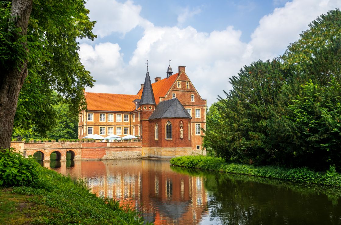 Das Schloss in Münster zwischen Bäumen und einem Fluss.