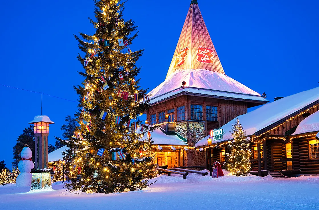 Joulupukin Pajakylä Rovaniemellä talvi-illassa, suuri jouluvalaistu kuusi ja luminen mökki Santa Claus Village -alueella.
